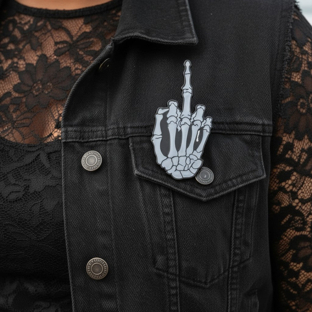 Mellow Monkey skeleton middle finger enamel pin with glow-in-the-dark effect pinned to a black denim vest.