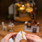 Hands assembling white cardboard pieces for a Mellow Monkey Slow Life Café DIY miniature house kit on a wooden table.