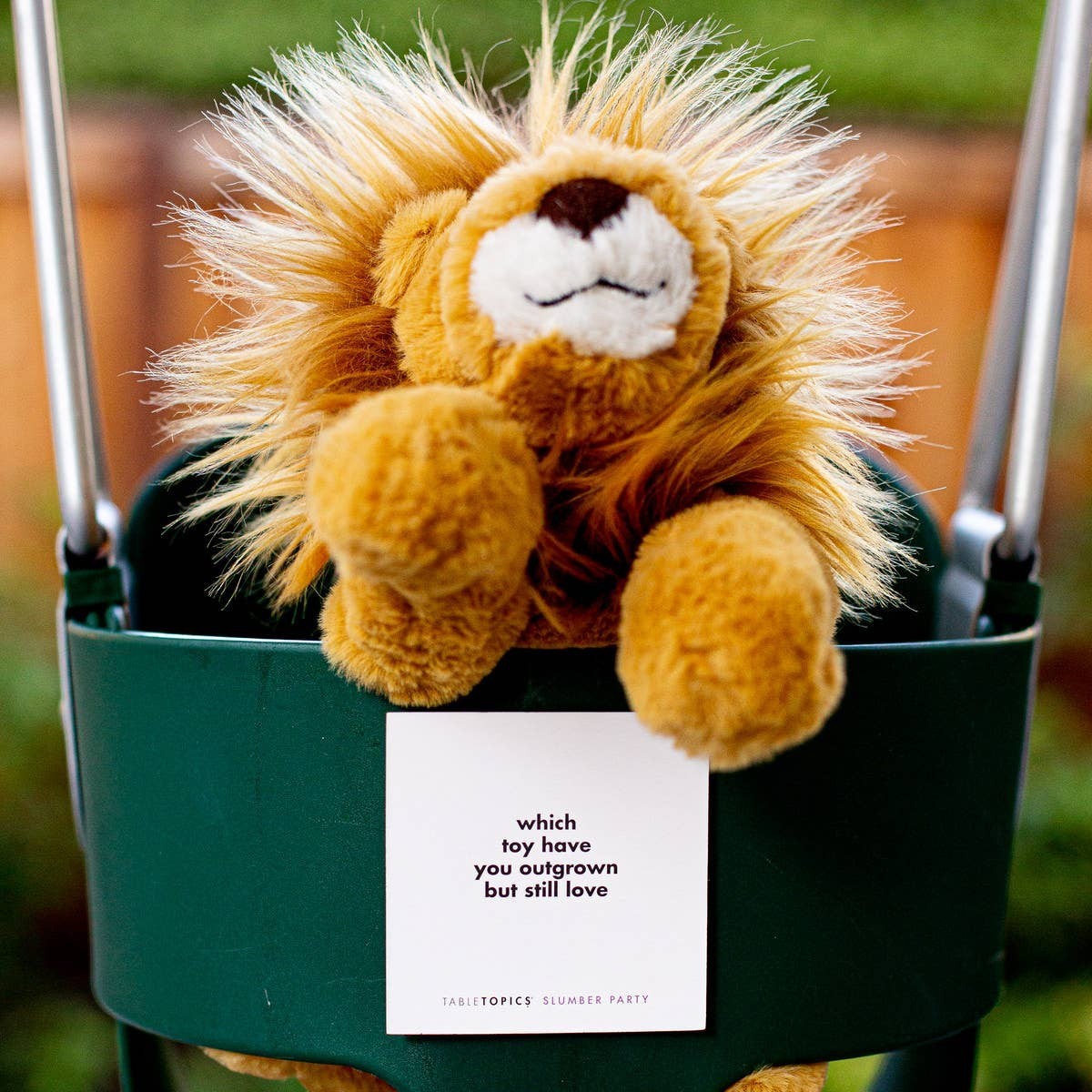 Mellow Monkey TABLETOPICS Slumber Party conversation card held by a plush lion toy in a green swing