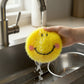 Mellow Monkey yellow smiley face dish scrubber being rinsed under a kitchen faucet