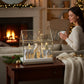 Mellow Monkey Snow City Lighted Terrarium with festive LED houses and trees on a wooden table by a fireplace.