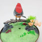Mellow Monkey sound-activated red bird toy perched on a branch with artificial flowers inside a decorative cage