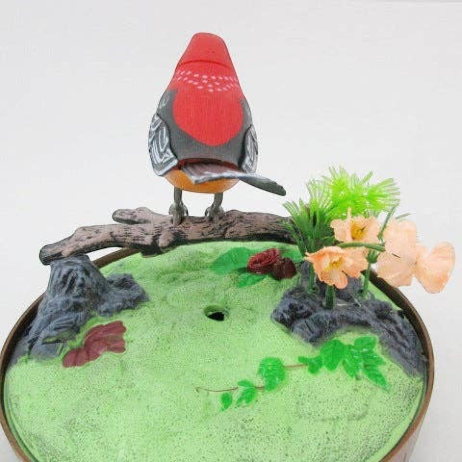 Mellow Monkey sound-activated red bird toy perched on a branch with artificial flowers inside a decorative cage