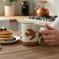 Mellow Monkey speckled stoneware mug with a hand-carved bird design held by a person next to a plate of pancakes.