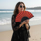 Woman in black lace dress holding a red and black Mellow Monkey spiderweb scallop fan on a sunny beach.