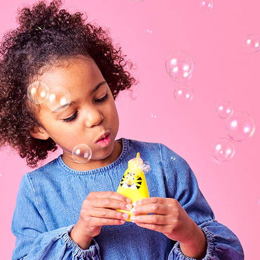 A young girl blowing bubbles with a Mellow Monkey Squeezy Bubbles yellow animal toy against a pink background.