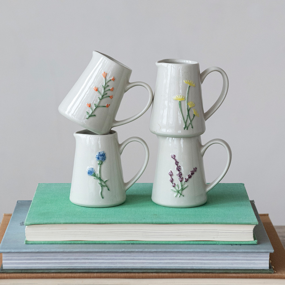 Mellow Monkey stoneware creamers with embossed botanical motifs in blue, yellow, purple, and orange, stacked on books.