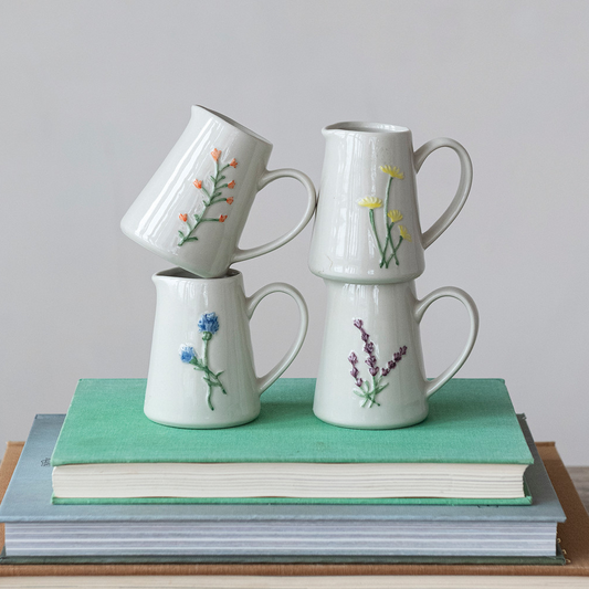 Mellow Monkey stoneware creamers with embossed botanical motifs in blue, yellow, purple, and orange, stacked on books.