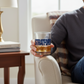 Mellow Monkey Stratford rocks glass filled with whiskey held by a person sitting in a cozy armchair