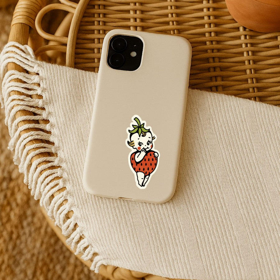 Mellow Monkey strawberry Kewpie sticker on a beige phone case resting on a textured cream cloth and rattan surface.