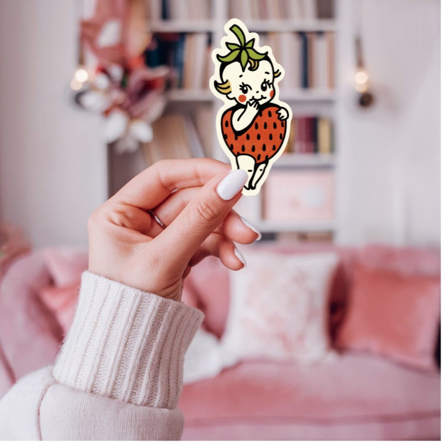Mellow Monkey vintage-inspired Strawberry Kewpie vinyl sticker held in a hand against a blurred pink living room