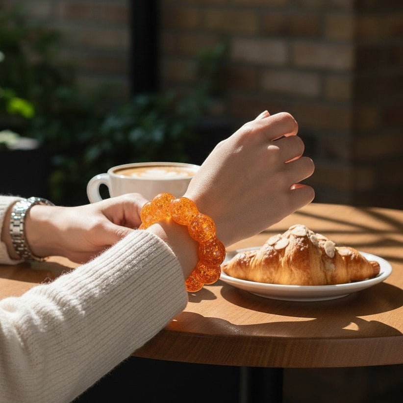 Mellow Monkey Sunburst Orange Feelzies Original sensory bracelet worn on a wrist at a cafe table with a croissant.