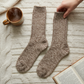 Mellow Monkey taupe ragg wool crew socks with a ribbed texture, laid on a cream cable knit blanket with a book and mug.