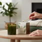 Mellow Monkey Thymes Frasier Fir candle in a green glass jar being lit on a wooden table with a cozy sweater.