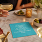 Mellow Monkey turquoise cocktail napkin with white text about living a comfortable life on a marble table setting