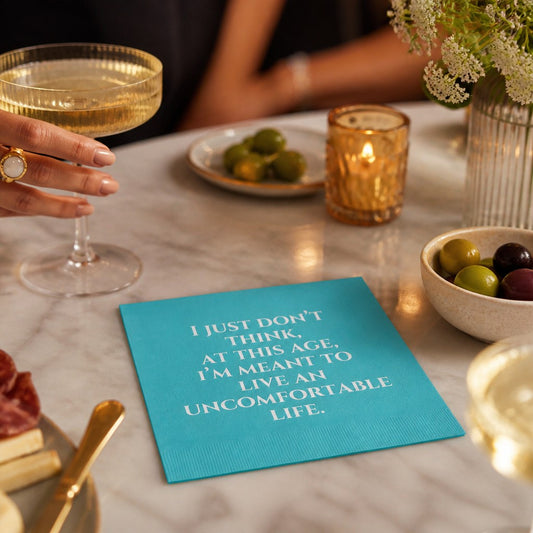 Mellow Monkey turquoise cocktail napkin with white text about living a comfortable life on a marble table setting