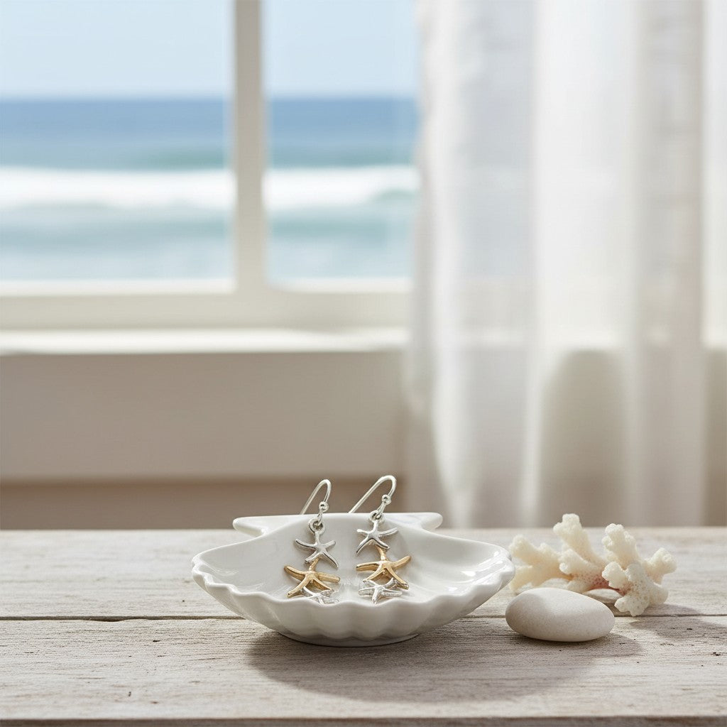 Mellow Monkey two-tone linked starfish earrings in a white shell dish on a wooden table with ocean view background.
