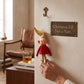 Mellow Monkey vintage-style elf ornament with a gold hat and red outfit hanging on a wall hook next to a sign.