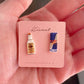 Mellow Monkey handmade vodka bottle and energy drink can stud earrings on a pink display card held in a palm.