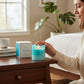 Mellow Monkey Volcano Artisan candle in a turquoise sea glass finish jar with a lit flame and smoke, next to its box.