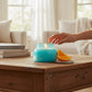 Mellow Monkey Volcano Artisan candle in a turquoise glass jar with a textured sea glass finish, lit flame, and orange slices on a wooden coffee table.