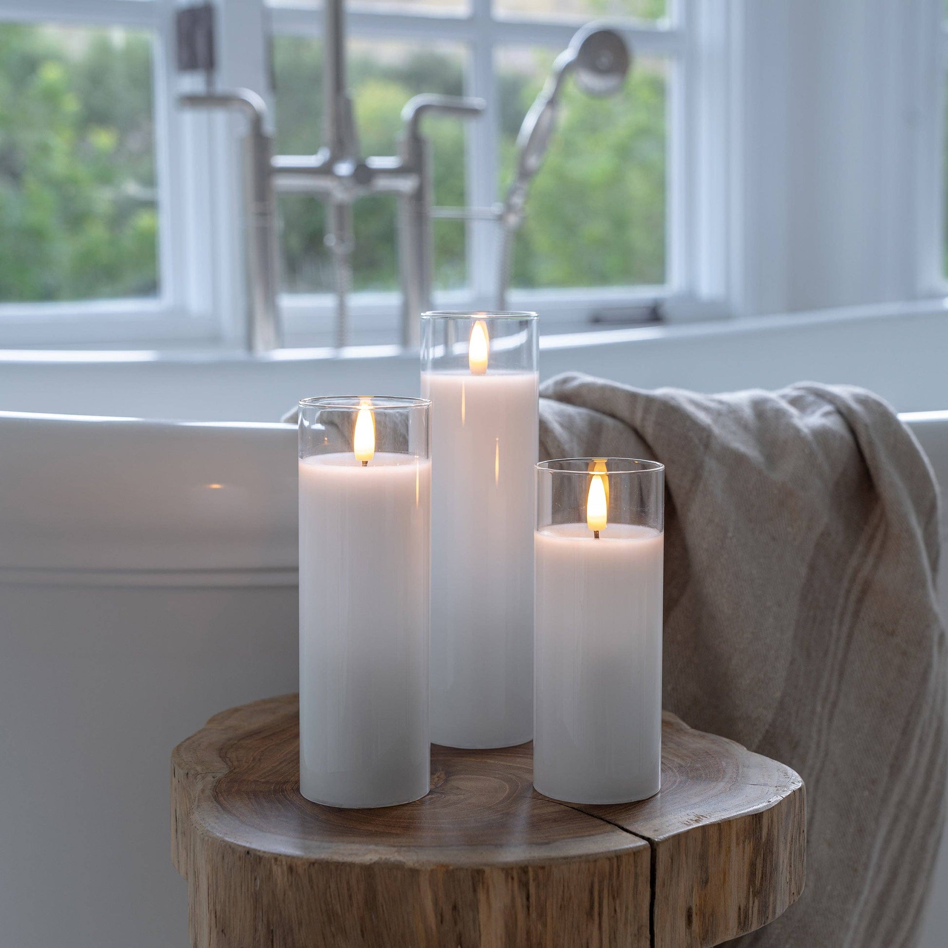 Mellow Monkey set of three white flameless LED pillar candles in glass holders on a rustic wooden stool in a bathroom.