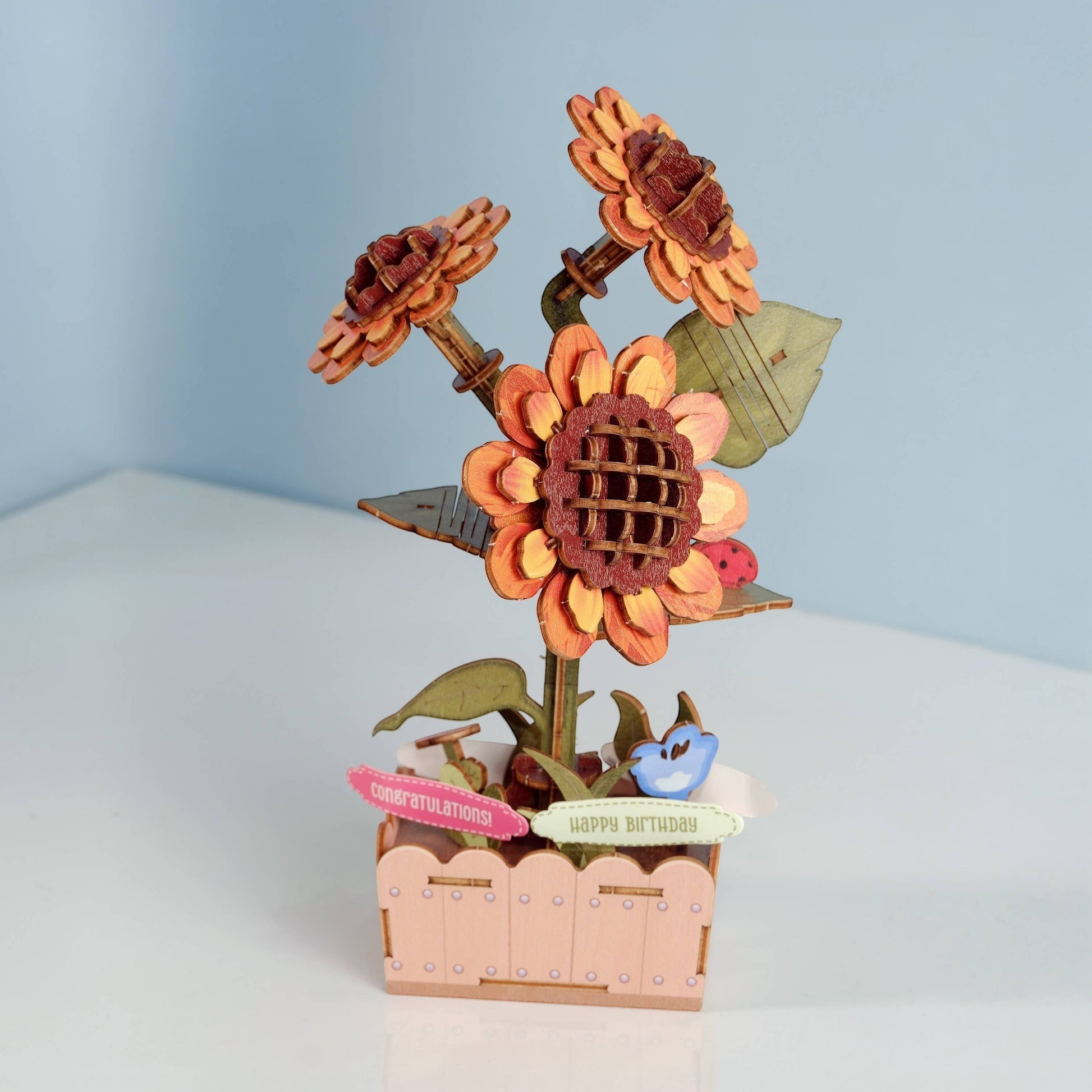 Mellow Monkey DIY wooden sunflower puzzle kit with orange and brown petals, green leaves, and a pink planter. Features "Happy Birthday" and "Congratulations" signs.