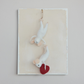 Mellow Monkey handcrafted white wool felt mice ornaments with a red heart, hanging on a textured cream board.
