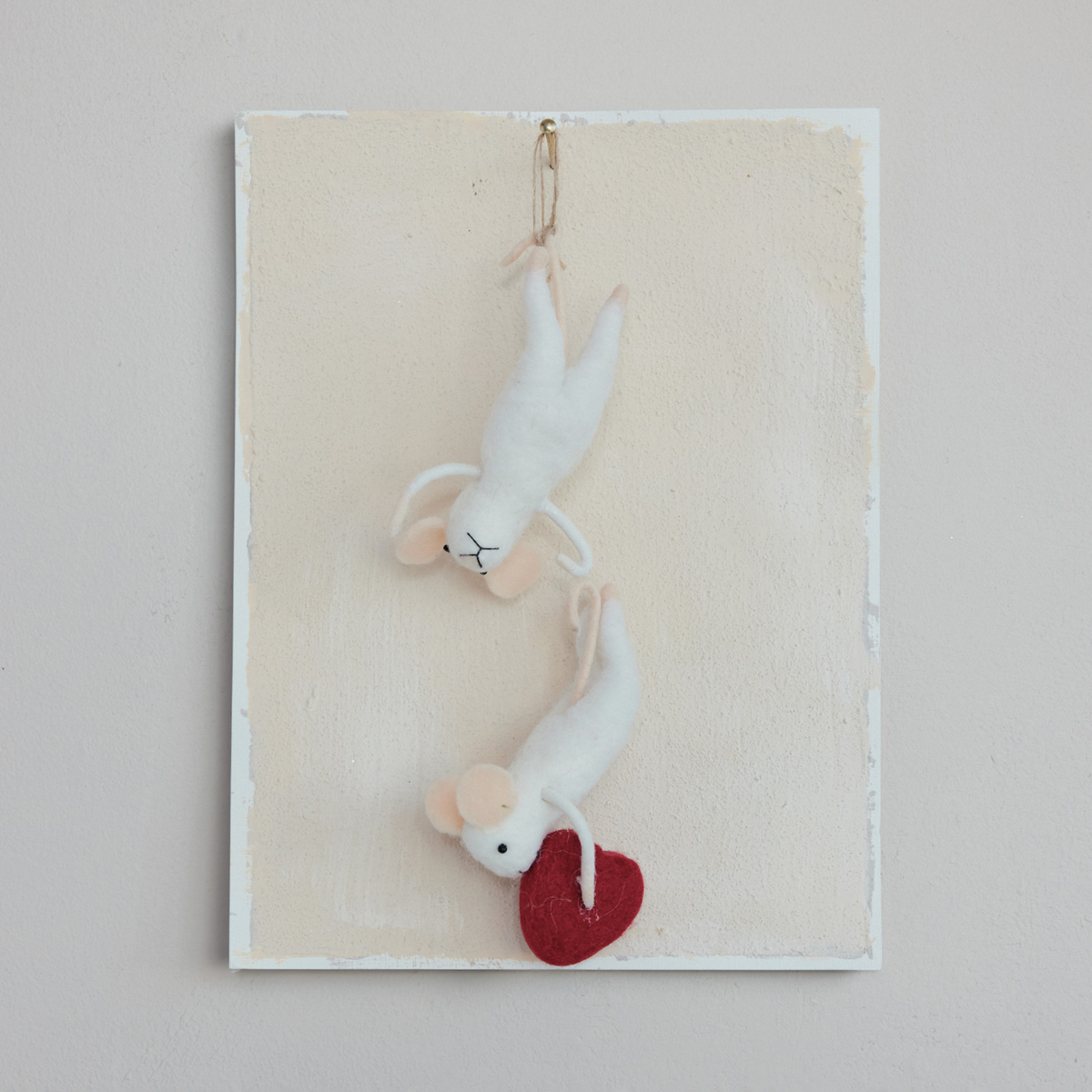 Mellow Monkey handcrafted white wool felt mice ornaments with a red heart, hanging on a textured cream board.