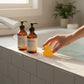 Mellow Monkey yellow rubber duck toy sitting on a tiled bathroom ledge next to shampoo bottles and a hand