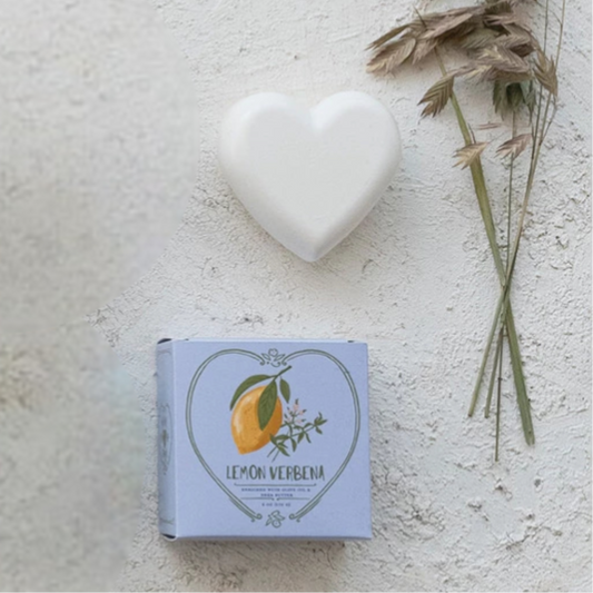 Heart-shaped soap on a textured surface with a box labeled 'Lemon Verbena' and dried herbs.